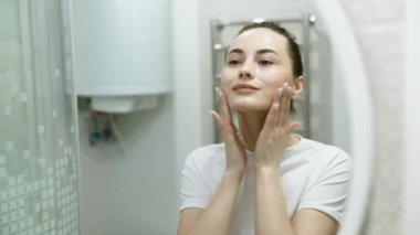 Güzel yüzlü ve mükemmel ciltli bir kadının ağır çekimleri kirlilikten arınıp gündüz veya gece kremine hazırlanmak için parmaklarıyla nazikçe şımartması..