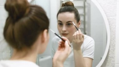Göz makyajına rimel süren mutlu genç bir kadın sabah hazırlanıyor, gülümsüyor makyaj yapıyor makyaj yapıyor, makyaj yapıyor evdeki banyoda aynaya bakıyor.