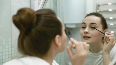 Göz makyajına rimel süren mutlu genç bir kadın sabah hazırlanıyor, gülümsüyor makyaj yapıyor makyaj yapıyor, makyaj yapıyor evdeki banyoda aynaya bakıyor.