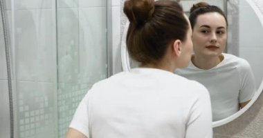 Krem sürdükten sonra güzel yüzlere dokunan mutlu çekici genç kadın aynaya bakıp doğal güzelliğin ve sağlıklı nemlendirilmiş cilt bakımının keyfini çıkar.