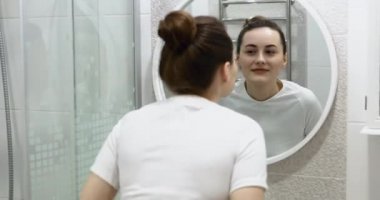 Krem sürdükten sonra güzel yüzlere dokunan mutlu çekici genç kadın aynaya bakıp doğal güzelliğin ve sağlıklı nemlendirilmiş cilt bakımının keyfini çıkar.