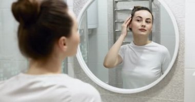 Krem sürdükten sonra güzel yüzlere dokunan mutlu çekici genç kadın aynaya bakıp doğal güzelliğin ve sağlıklı nemlendirilmiş cilt bakımının keyfini çıkar.