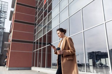 Güneş gözlüklü, gülümseyen kıvırcık kadın Central City caddesinde yürüyor ve telefonunu kullanıyor. Kahverengi ceketli güzel yaz kadını caddede yürürken cep telefonuna bakıyor.