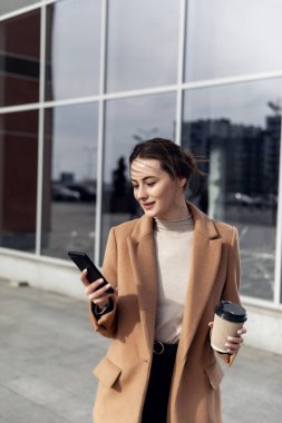 Güneş gözlüklü, gülümseyen kıvırcık kadın Central City caddesinde yürüyor ve telefonunu kullanıyor. Kahverengi ceketli güzel yaz kadını caddede yürürken cep telefonuna bakıyor.