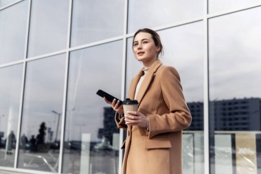Akıllı telefonlu genç bir Cousian kadın caddenin karşısında duruyor. Bina geçmişi bulanık. Siyah süitte, bir fincan kahve ve telefonla güzel bir kızın moda fotoğrafı.