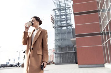 Moda kıyafetli şık bir kadın şehirde yürüyor, cep telefonu kullanıyor, kahve içiyor. Moda tarzı olan çekici bir kadın. Kafasında siyah şapka vardı. Kağıt bardağı içkiyle tutmak