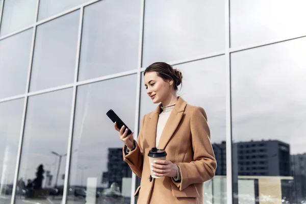 Güneş gözlüklü, gülümseyen kıvırcık kadın Central City caddesinde yürüyor ve telefonunu kullanıyor. Kahverengi ceketli güzel yaz kadını caddede yürürken cep telefonuna bakıyor.