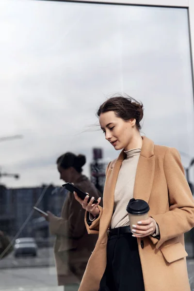 Smiling curly woman wearing trendy sunglasses walks down the central city street and uses her phone. Pretty summer woman in brown jacket walks down the street looking at her mobile phone