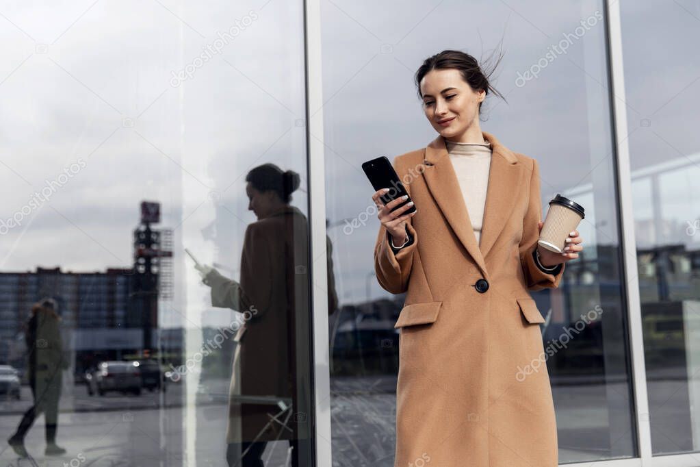 Smiling curly woman wearing trendy sunglasses walks down the central city street and uses her phone. Pretty summer woman in brown jacket walks down the street looking at her mobile phone