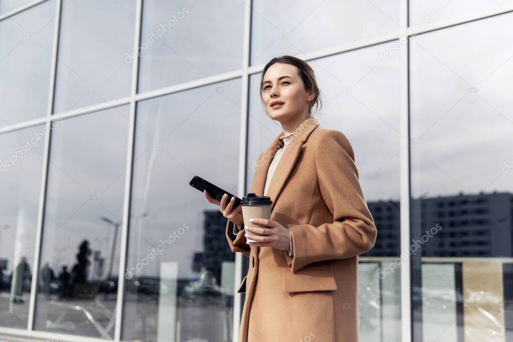 Young cousian woman with smartphone standing against street blurred building background. Fashion business photo of beautiful girl in black casual suite with phone and cup of coffee