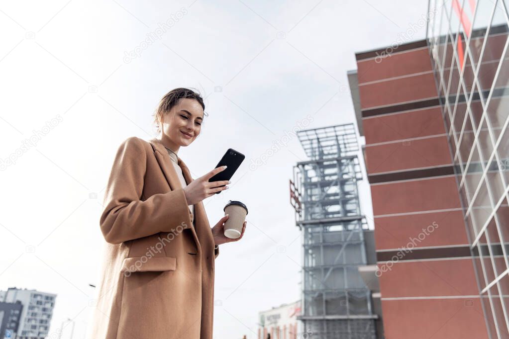Smiling curly woman wearing trendy sunglasses walks down the central city street and uses her phone. Pretty summer woman in brown jacket walks down the street looking at her mobile phone