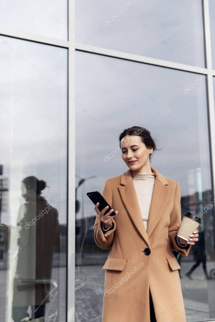 Business Woman smiling charmingly, using her Smartphone. Female enjoying sunny day and funny conversation. Typing on her Phone with Interest. Brunette Girl. Successful Woman. Phones. Apps.