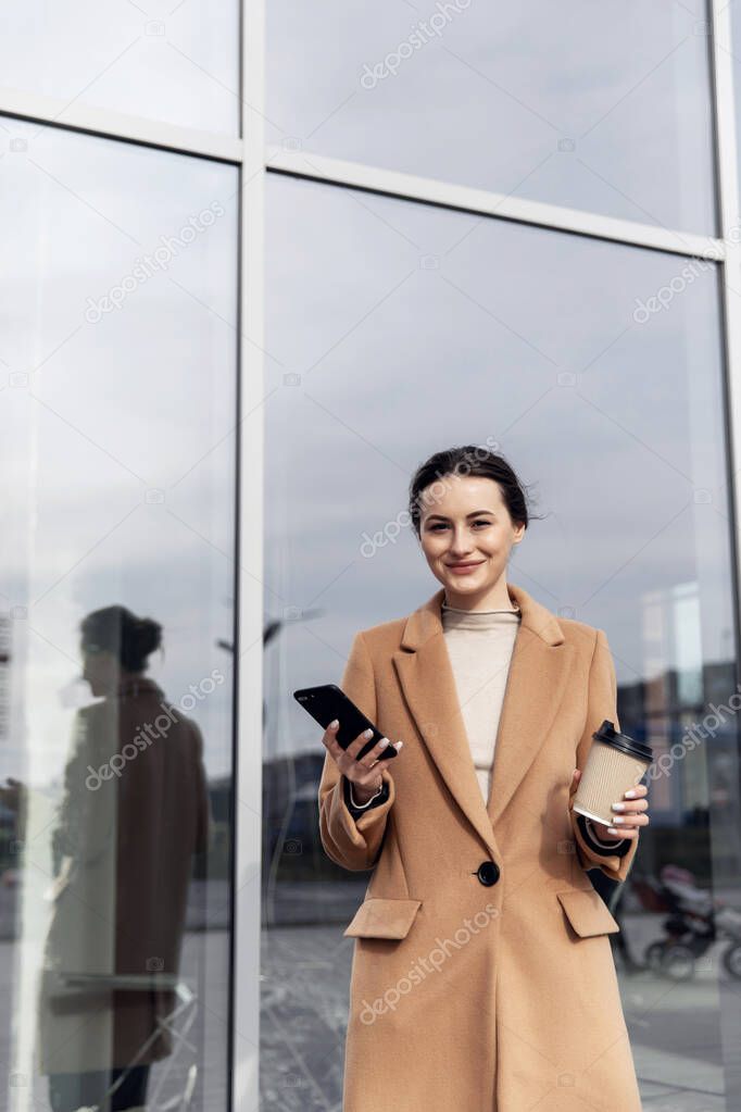 Closeup young Business Woman holding smartphone, Coffee to go and smiling. Attractive stylish Woman using her Phone near modern Building. Having Break. Smartphones. Games. People. Emotions. Business.