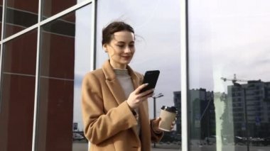 Akıllı telefonlu genç bir Cousian kadın caddenin karşısında duruyor. Bina geçmişi bulanık. Siyah süitte, bir fincan kahve ve telefonla güzel bir kızın moda fotoğrafı.