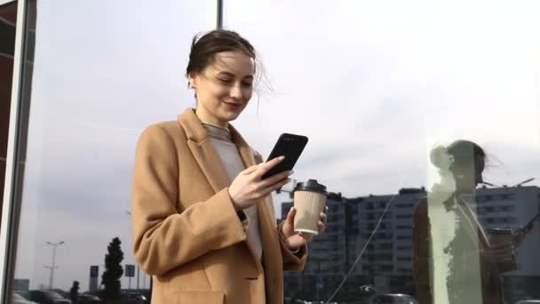 年轻的库西亚女人拿着智能手机站在街道上模糊的建筑背景下 穿着黑色休闲装 手持电话和咖啡的美女的时尚商业照片 — 图库视频影像