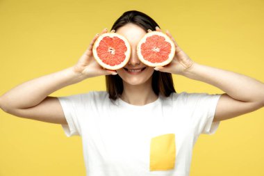 Kareli tişörtlü, komik, genç bir kadın. Gözlerine turunçgillerden yarımlar tutuyor ve sarı arka planda ördek suratı yapıyor.