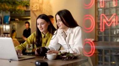 Gündelik kıyafetli iki genç esmer restoranda dizüstü bilgisayardan alışveriş yapıyor. Gülümseyen bayan arkadaşlar birbirlerine uygun kıyafetler seçmede yardım ediyorlar..