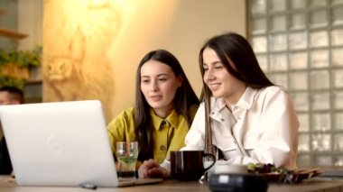 Uzun kahverengi saçlı, modaya uygun kıyafetleri olan güzel kadınlar rahat bir kafede kişisel bilgisayarlarıyla ders çalışıyorlar. Ders hazırlamak için internet üzerinden bilgi arayan kız öğrenciler.