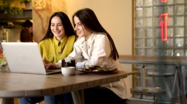 Parlak elbiseli iki bayan arkadaş, rahat bir kafede otururken dizüstü bilgisayardan uçak bileti alıyorlar. Kahverengi saçlı mutlu kadınlar yaz tatili için yeni bir gezi planlıyorlar..
