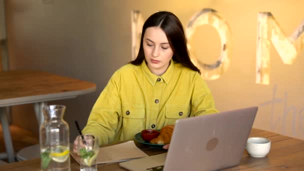 Sarı tişörtlü, kendine güvenen bir bayan öğrenci kişisel laptopuyla masada oturuyor ve not alıyor. Kahverengi saçlı genç kız, ödev yaparken kahve dükkanında vakit geçiriyor..