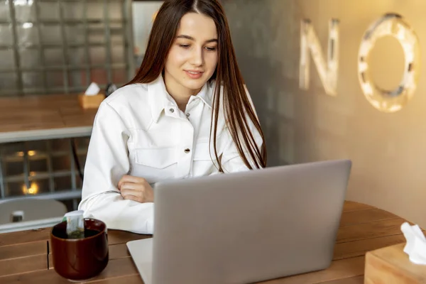 Beautiful brunette in white trendy shirt sitting at table and browsing internet on personal laptop. Young girl using wifi at modern cafe for working on distance