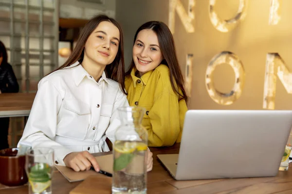 Cheerful young ladies in stylish clothing hugging each other after choosing new destination for travelling. Beautiful ladies with brown hair using laptop for buying tickets online.