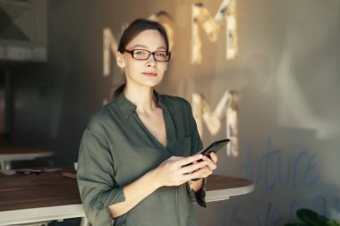 Süslü gözlüklü güzel bir kadın elinde kağıt bardakla kafeteryada otururken akıllı telefona bakıyor. Cep telefonu kullanan mutlu bir kadın. İş kadını kahve içiyor ve gülümsüyor..