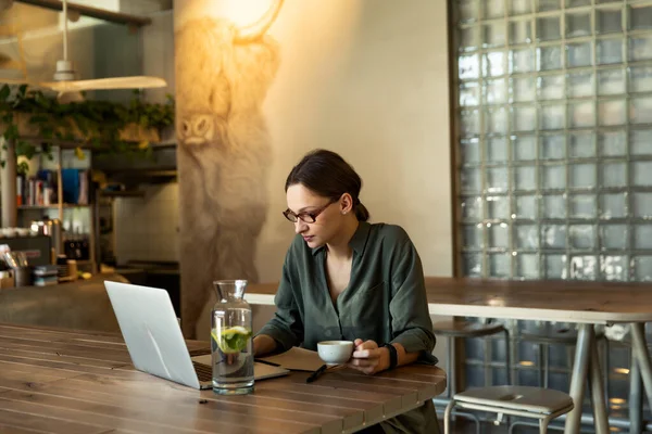 Woman in stylish glasses and black jacket drinking coffee at a restaurant and writing text on mobile laptop