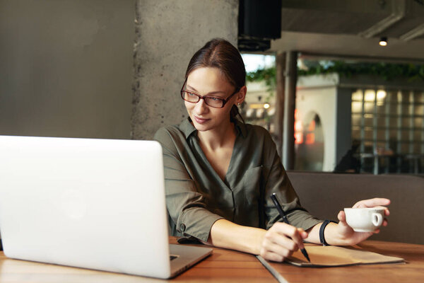 Деловая женщина в черном костюме и очках сидит за столом в кафе, работая за ноутбуком, держа в руках ноутбук с записями
