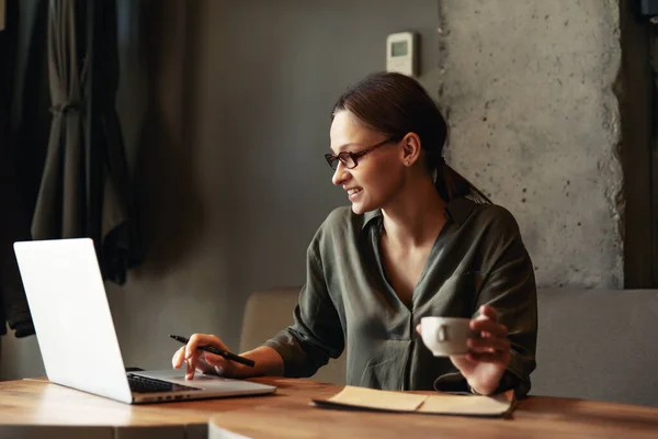 Focused business woman in stylish glasses sit on cafe working on laptop, concentrated serious female working with computer and cappuccino in coffee shop, freelancer, studying online, browse internet, checking bills