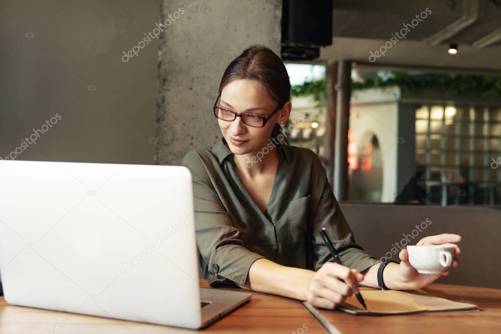 Business woman in a black suit and glasses sitting at a table in a cafe working at a laptop holding a notebook with records