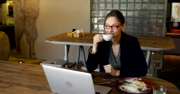 スタイリッシュなメガネと黒のジャケットを着た女性がレストランでコーヒーを飲み 携帯電話でテキストを書きます — ストック動画