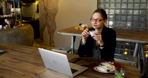 スタイリッシュなメガネと黒のジャケットを着た女性がレストランでコーヒーを飲み 携帯電話でテキストを書きます — ストック動画