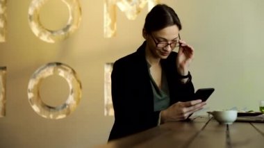 Şık gözlüklü ve siyah ceketli bir kadın restoranda kahve içiyor ve cep telefonuyla mesaj yazıyor.