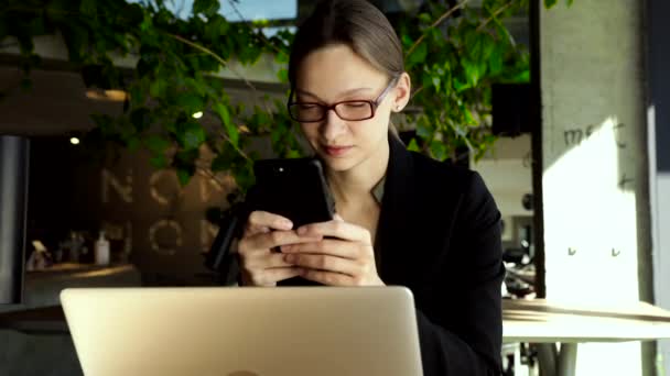 漂亮的女人戴着时髦的眼镜 手里拿着咖啡杯 坐在自助餐厅里看着智能手机 快乐的女人用手机 女商人一边喝咖啡一边笑 — 图库视频影像