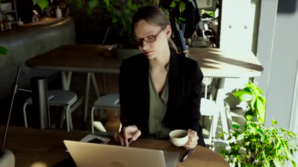 戴着时髦眼镜的漂亮的女商人坐在室内咖啡桌旁 在笔记本电脑上工作 看着相机 — 图库视频影像