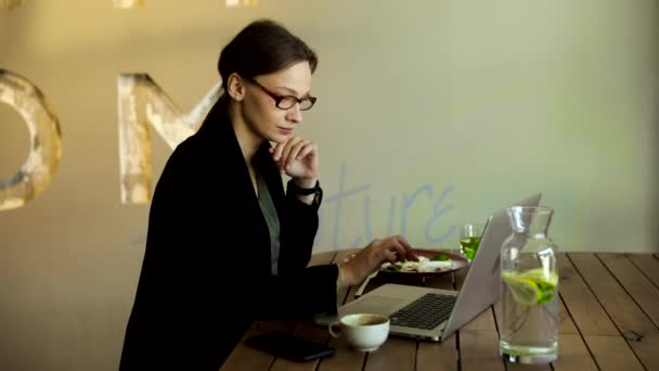 戴着时髦眼镜的商界女性专心致志地坐在咖啡店的笔记本电脑上 专心致志地与电脑和卡布奇诺一起工作的女性 自由职业者 在线学习 浏览互联网 核对账单 — 图库视频影像