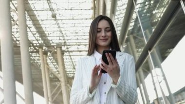 Güneş gözlüklü, gülümseyen kıvırcık kadın Central City caddesinde yürüyor ve telefonunu kullanıyor. Beyaz ceketli güzel yaz kadını caddede yürürken cep telefonuna bakıyor.