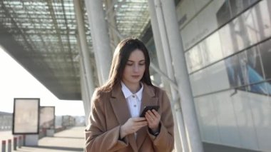 Business Woman havalimanı yakınlarında akıllı telefonunu kullanarak büyüleyici bir şekilde gülümsüyor. Kadın güneşli günün ve komik sohbetin tadını çıkarıyor. Telefonuna faiziyle yazı yazıyormuş..
