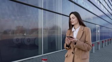 Güneş gözlüklü, gülümseyen kıvırcık kadın Central City caddesinde yürüyor ve telefonunu kullanıyor. Beyaz ceketli güzel yaz kadını caddede yürürken cep telefonuna bakıyor.