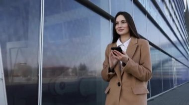 Güneş gözlüklü, gülümseyen kıvırcık kadın Central City caddesinde yürüyor ve telefonunu kullanıyor. Beyaz ceketli güzel yaz kadını caddede yürürken cep telefonuna bakıyor.