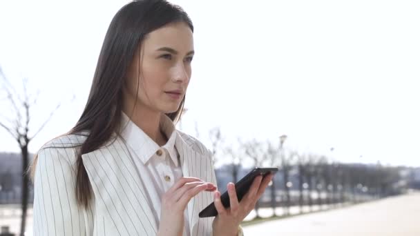 美丽的年轻女商人穿着白衬衫和步行在城市 在外面 慢动作手机上键入文本消息的专业女雇主伸长时使用现代的智能手机 — 图库视频影像