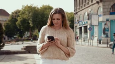 Mavi beyaz çizgili tişört giyen gülümseyen genç bir kadın akıllı telefon kullanarak eski caddede yürüyor. İletişim, sosyal ağlar, online alışveriş kavramı.