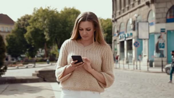 青と白のストライプのドレスシャツを着た若い女性がスマートフォンを使って古い通りを歩いているのを笑っています。コミュニケーション、ソーシャルネットワーク、オンラインショッピングの概念. — ストック動画