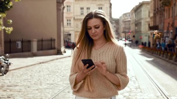 Mulher bonita com encantador sorriso andando na rua e usando telefone celular. Tendo óculos de sol modernos e vestindo roupas elegantes. Casualmente vestido Jovem mulher atraente — Vídeo de Stock