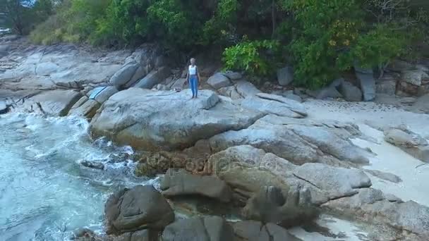 AERIAL. Mujer joven de pie sobre las rocas de la costa oceánica en los trópicos . — Vídeos de Stock