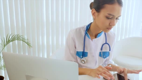 Γιατρός, πληκτρολογώντας κείμενο σε ένα φορητό υπολογιστή. Νεαρά ασιατικές ελκυστική γυναίκα γιατρός κάθεται στο γραφείο στο τραπέζι. Ασπίδα με το λογότυπό σας σε ένα φορητό υπολογιστή — Αρχείο Βίντεο