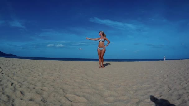 Sesión de fotos en la playa. Una mujer joven, esbelta, bronceada, imitando un intento de detener un avión de vuelo bajo con un gesto de autostop . — Vídeo de stock