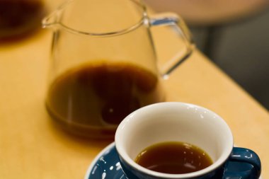 Kahve dükkanında fincan tabağı olan mavi seramik fincan, sert, siyah, canlandırıcı bir kahveyle ahşap masanın üzerinde. Amerikalı. Sabah içkisi. Sürahi. Ware..