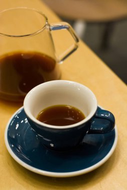 Kahve dükkanında fincan tabağı olan mavi seramik fincan, sert, siyah, canlandırıcı bir kahveyle ahşap masanın üzerinde. Amerikalı. Sabah içkisi. Sürahi. Ware..
